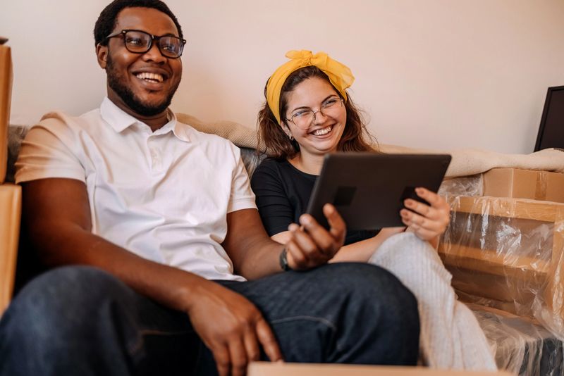 Young couple using digital tablet while moving in new apartment and surrounded with boxes