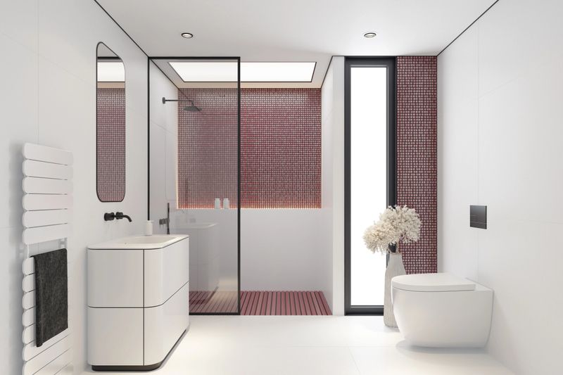 Contemporary bathroom interior featuring a clean, minimalist design. Red and white. The space includes a white vanity with a built-in sink and a single drawer, paired with a rounded-edge mirror and matte black wall-mounted faucets. A white towel radiator with a dark gray towel adds contrast on the left, while a glass-enclosed shower with black fixtures and a handheld attachment anchors the right side. The shower floor is designed with white slats, and the back wall showcases a mosaic tile accent. Neutral tones and modern finishes create a serene, functional atmosphere ideal for luxury residential or hospitality settings.