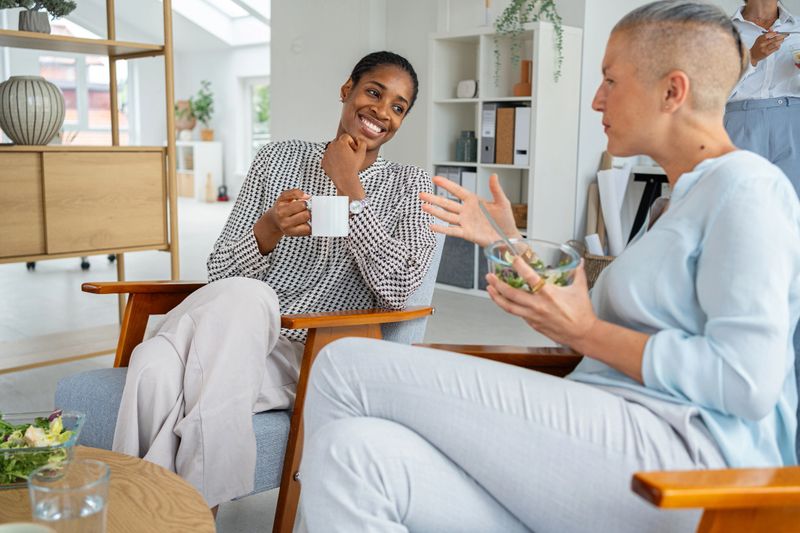Two women engage in conversation while having a light meal in a modern workspace, enjoying a relaxed and friendly environment. The image conveys themes of communication, casual dining, and camaraderie.