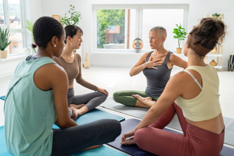 A group of four diverse women sitting cross-legged in a yoga studio, engaging in a peaceful and focused discussion as part of their yoga session. The room is bright and serene.