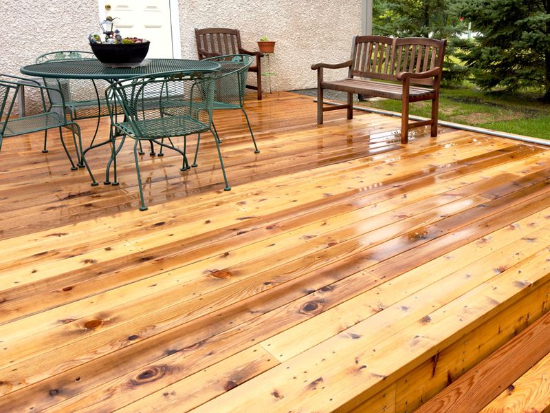 freshly washed wooden patio deck with green metal table set, wooden bench, and potted plants, surrounded by lush greenery and house exterior