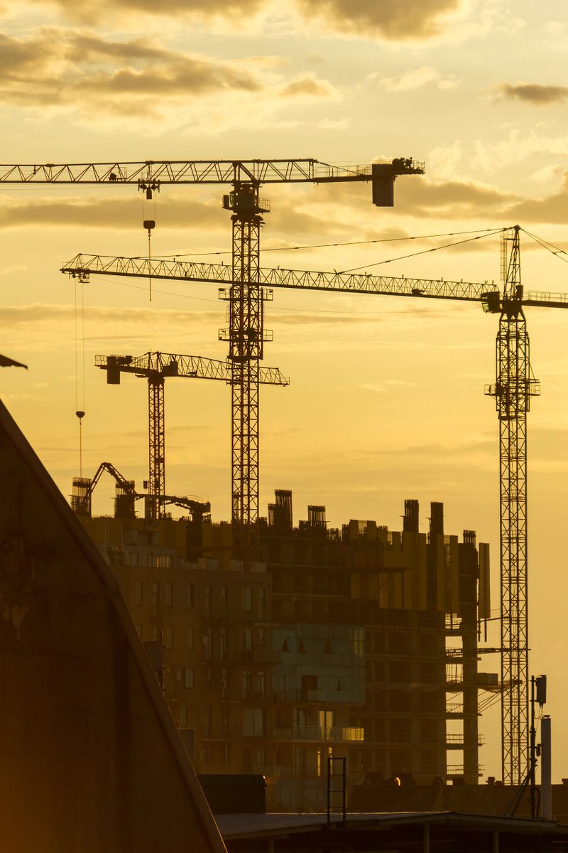 Atmospheric photo of tall construction cranes and an active construction site silhouetted against a colorful sunset sky. The image captures the spirit of progress, development, and urban transformation. Ideal for themes related to architecture, engineering, real estate growth, city planning, and industrial ambition.
