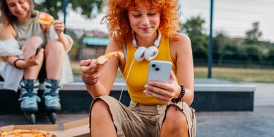 Two friends enjoying pizza outdoors, one checking her phone with headphones around her neck.