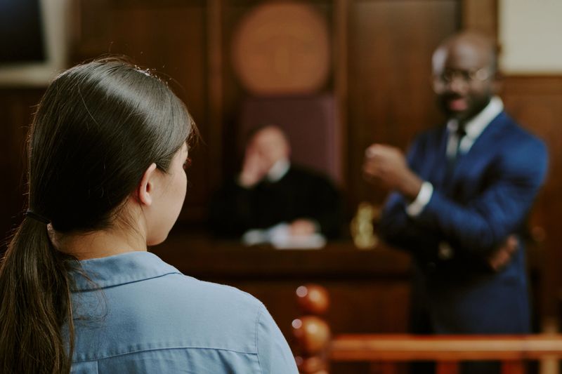 Caucasian young woman standing in courtroom facing Black middle aged man gesturing while judge sitting in background listening during legal proceeding or testimony