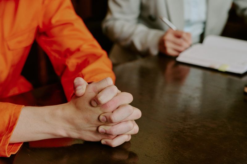 Caucasian young adult man sitting at table with hands clasped in front, wearing orange jumpsuit, legal professional writing in notebook during meeting or consultation
