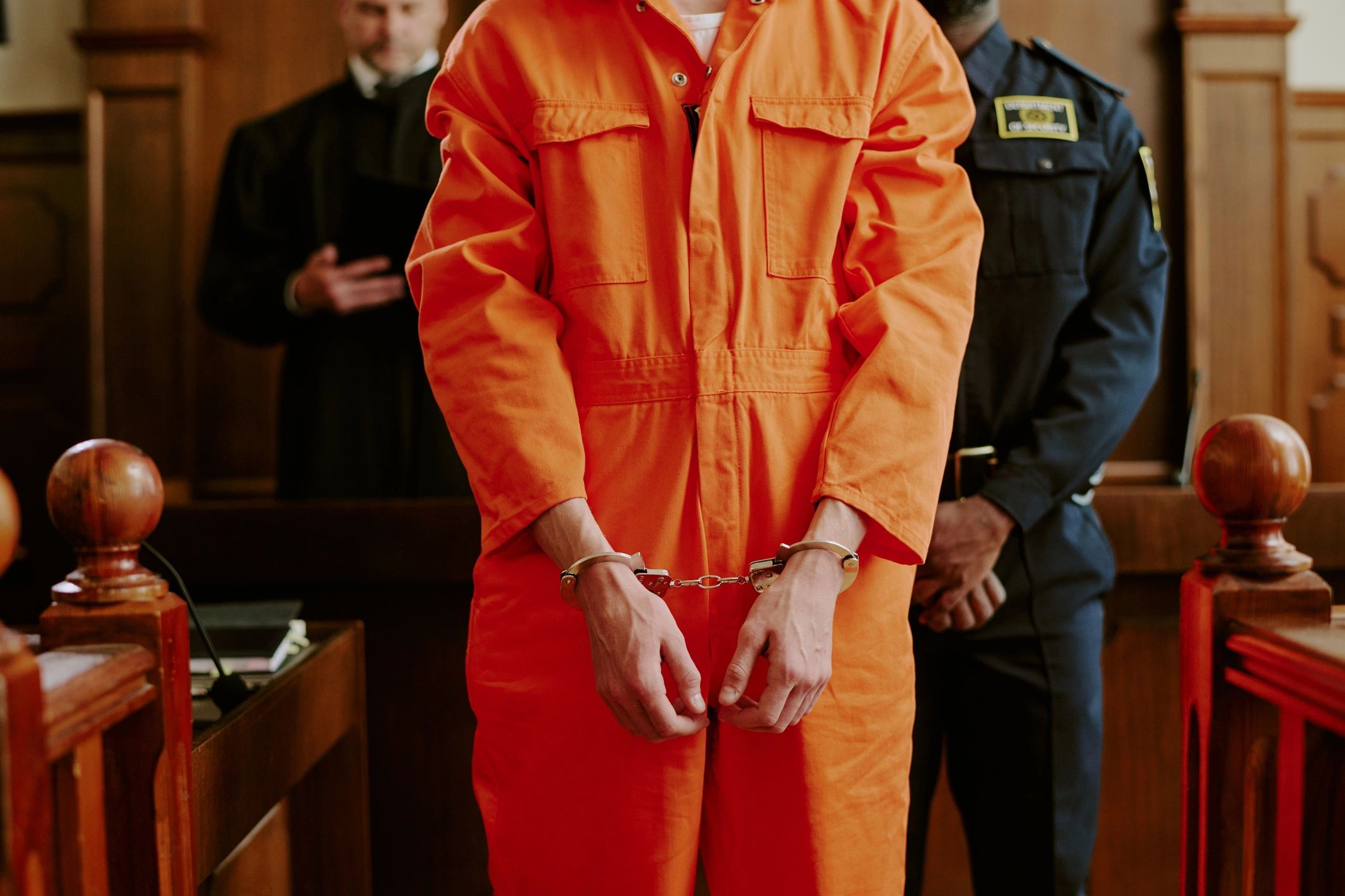 Inmate in orange jumpsuit stands handcuffed in court with guard and judge behind.