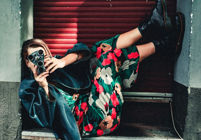 Dynamic urban portrait of a stylish young woman reclining casually in a doorway, aiming a vintage film camera at the viewer. Her vibrant floral trousers contrast with a dark oversized jacket and sturdy leather boots, blending retro aesthetics with modern street style. The vivid colors, playful composition, and confident pose create a cinematic and editorial feel — ideal for fashion features, lifestyle concepts, and creative storytelling.