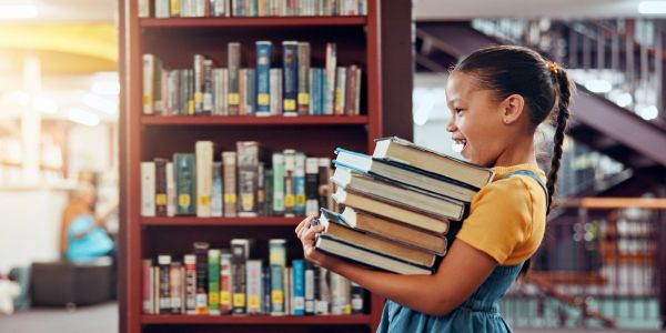 Young girl carrying a stack of books in a library.