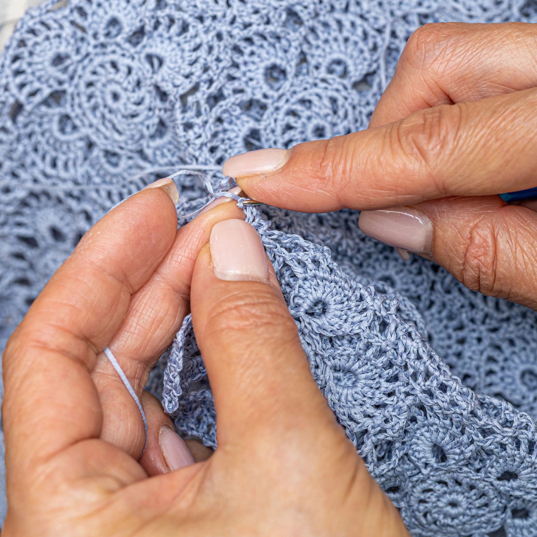 Hands crocheting intricate blue lace fabric with fine thread.