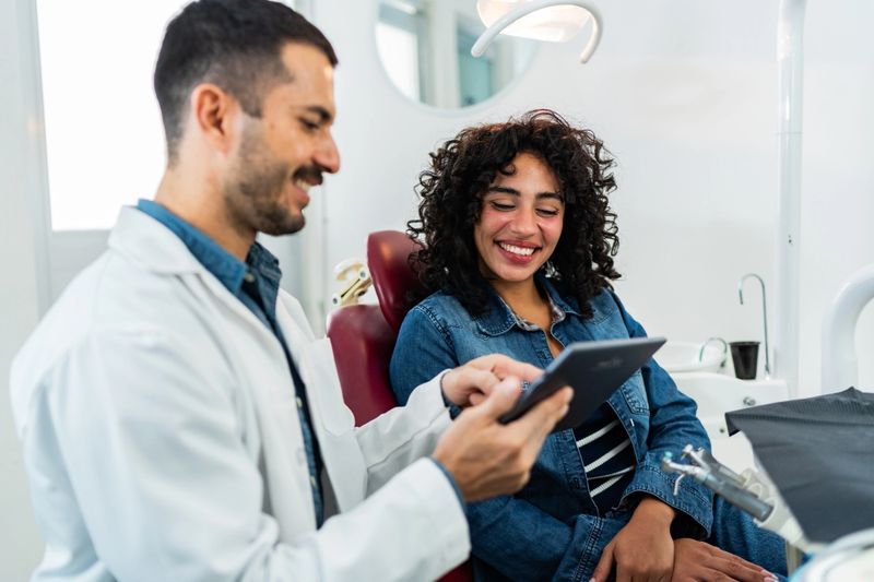 Mid adult dentist man using digital tablet and talking to young patient woman at dentist's office
