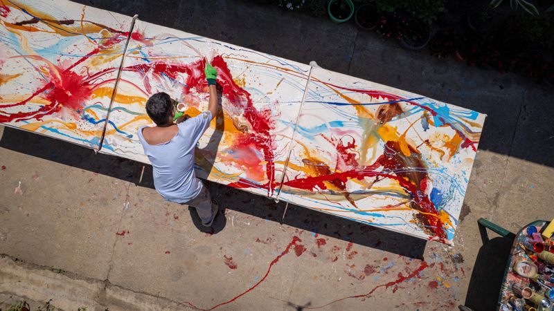 Shot from a drone from a high angle, a male artist is in the process of creating an abstract painting on a large canvas in an outdoor courtyard.