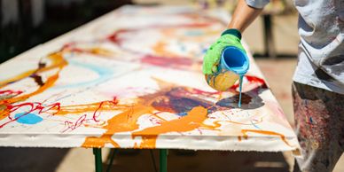 An artist pours blue paint onto a colorful abstract canvas outdoors.