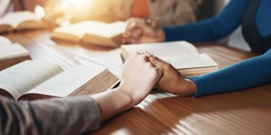 People holding hands over open books on a table.