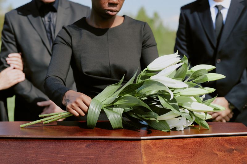 Black woman placing lilies on wooden casket surrounded by Caucasian men in formal attire during outdoor funeral ceremony, hands gently arranging flowers in remembrance