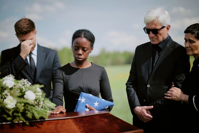 Black young adult woman holding folded American flag standing beside closed casket, with Caucasian middle aged man in sunglasses and diverse adults mourning outdoors during funeral