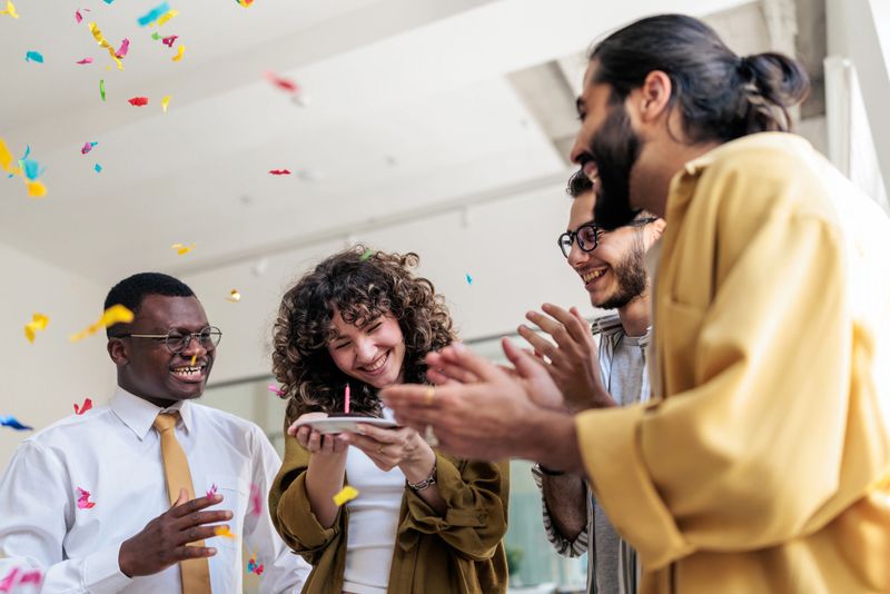 A group of coworkers celebrating a colleague’s birthday in the office with a small cake topped with a candle, party hats, and confetti, enjoying friendship and a cheerful atmosphere at the workplace.