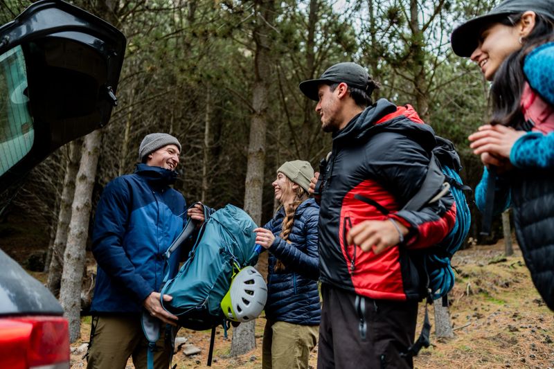 A group of friends organizes backpacks, ropes, and climbing gear next to the car before starting their adventure in the forest. Their expressions show enthusiasm and companionship at the beginning of the trip