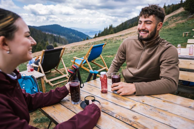 Two hikers relax in a mountaintop restaurant after a hike, drinking fresh juice and enjoying a stunning panoramic view of nature.