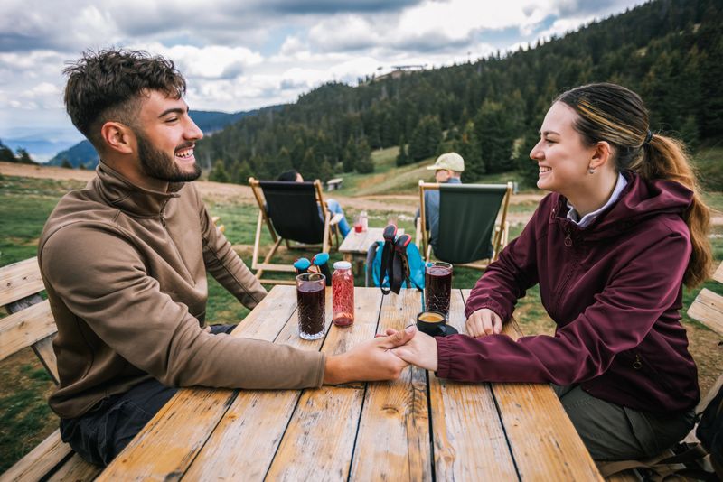 A romantic hiking couple sits in a mountaintop restaurant, holding hands while enjoying fresh juice and the stunning natural view.