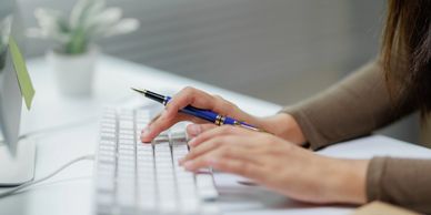 Person typing on a white keyboard with a blue pen in hand.