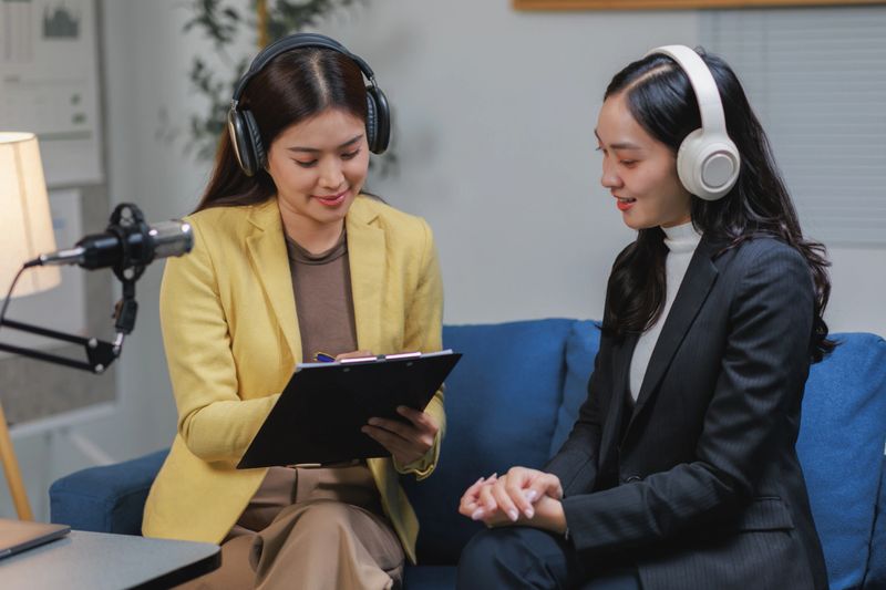 Two young women podcasters wearing headphones and taking notes during a recording session in a studio