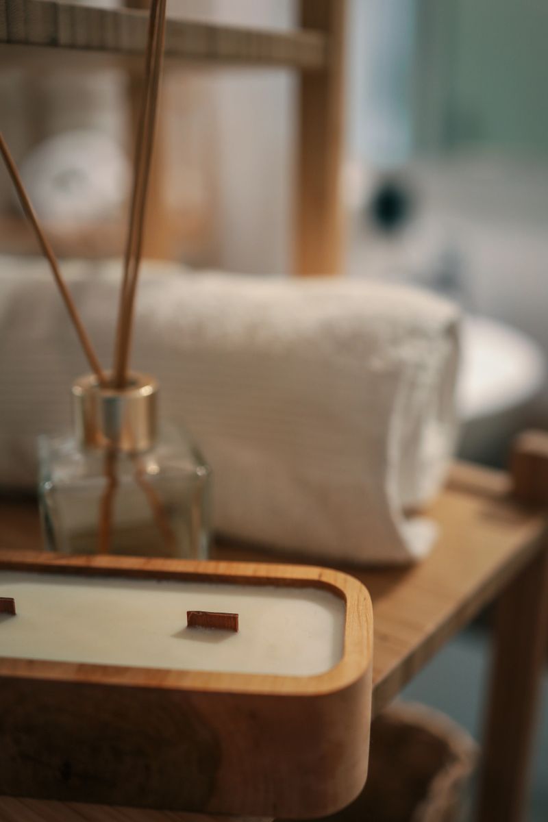 Close-up of a wooden candle, glass aroma diffuser, and rolled towel on a bamboo shelf in a cozy bathroom. Soft light and natural textures create a relaxing atmosphere.