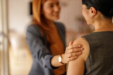 A woman placing a comforting hand on another's shoulder in a warm indoor setting.