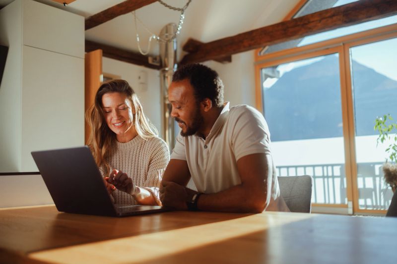A couple is sitting together at a wooden table in their living room, engaged in a collaborative task on a laptop, with the warm sunlight and a view of the mountains through the window creating a pleasant backdrop.