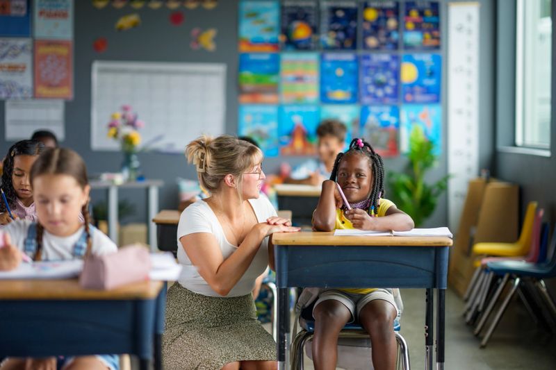 A teacher interacts warmly with students in a lively classroom environment, highlighting diversity and learning engagement. The background showcases colorful educational materials and a welcoming, dynamic atmosphere.