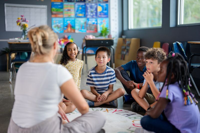 A teacher interacts with a group of students sitting together in a classroom. The scene highlights multicultural diversity and depicts a collaborative, learning-focused environment. Bright lighting and classroom decor enhance the setting.