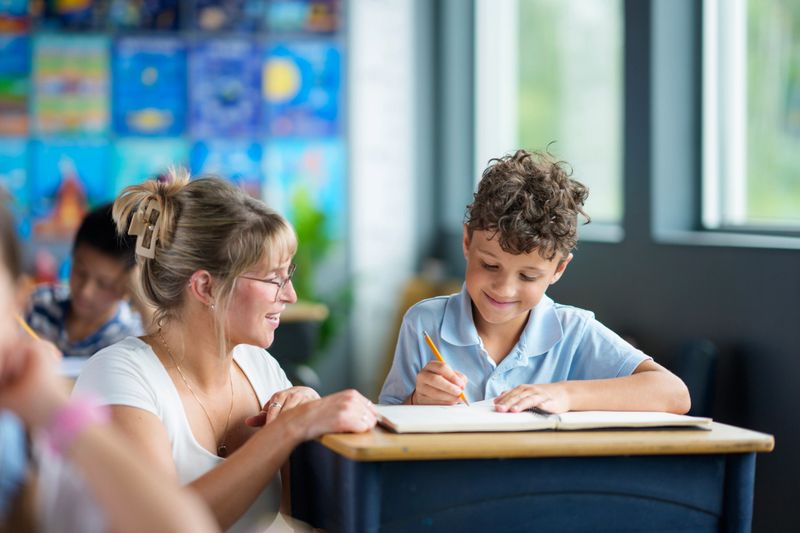 A caring teacher assists a young student in a brightly lit classroom, emphasizing learning and mentoring. The setting enhances focus, with a positive atmosphere fostering student-teacher interaction and educational growth.