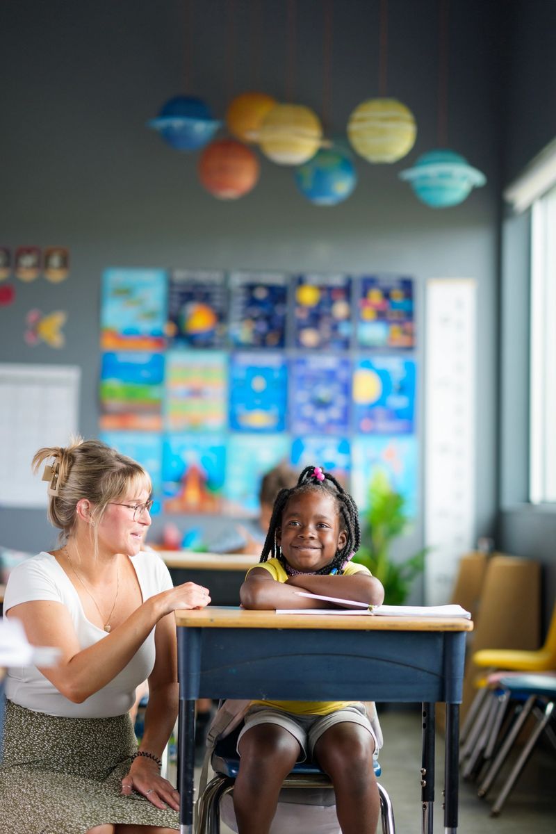 A teacher assists a student in a colorful classroom featuring a science theme with planets. The child appears excited and engaged, highlighting the positive atmosphere and diversity in educational settings.