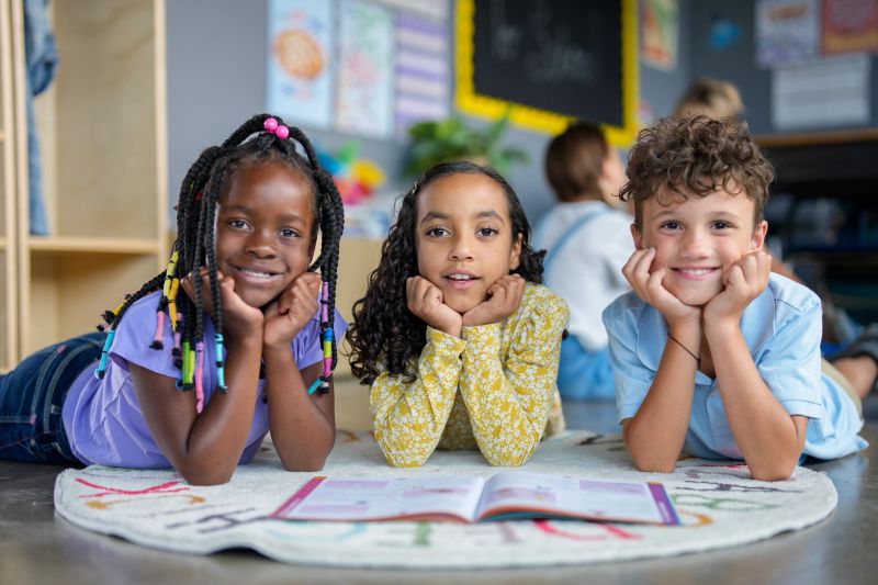 Three cheerful children in a classroom, highlighting cultural diversity and friendship. They read together, foster a warm educational environment, and exemplify classroom collaboration.
