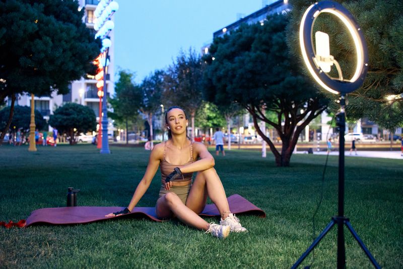 A fitness influencer conducts a workout session on a yoga mat outdoors with ring light setup.