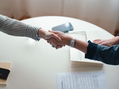 Two people shaking hands over a table with documents and phones.