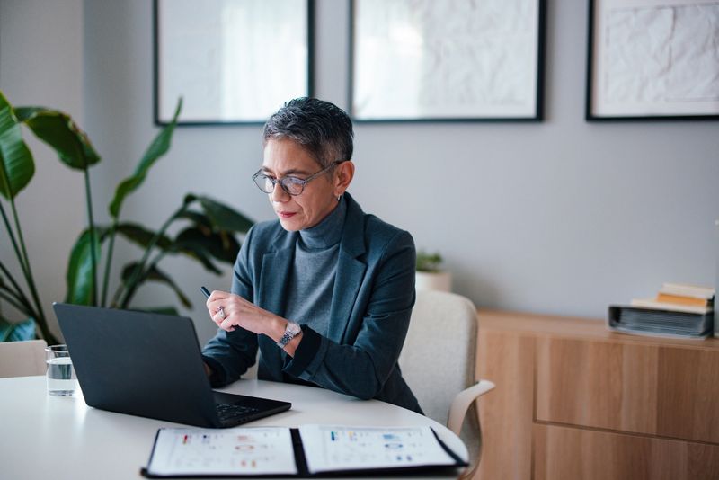 A businesswoman reviews documents while working on a laptop in her professional office. There are decorative plants and modern furnishings creating a contemporary and focused workplace atmosphere.