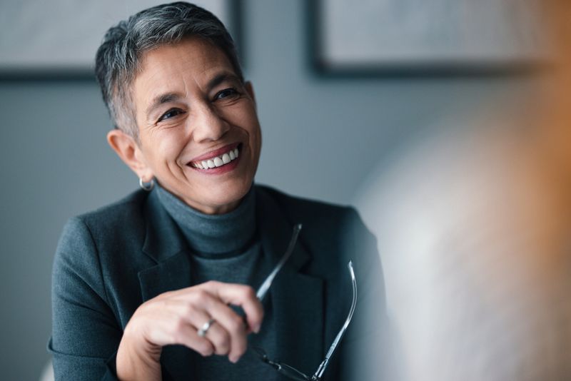A professional woman with short gray hair, smiling warmly while holding glasses during a conversation in a modern office environment. The setting conveys themes of confidence, communication, and leadership in a business context.