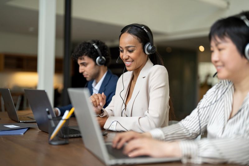 Three diverse coworkers wearing headsets working on laptops in an office, focusing on customer support and teamwork, expressing professionalism and collaboration in a corporate or business environment.