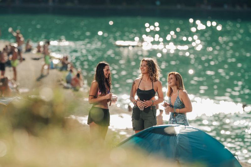 Three young women are laughing and enjoying drinks by a lake at a music festival, with a tent and other festivalgoers in the background
