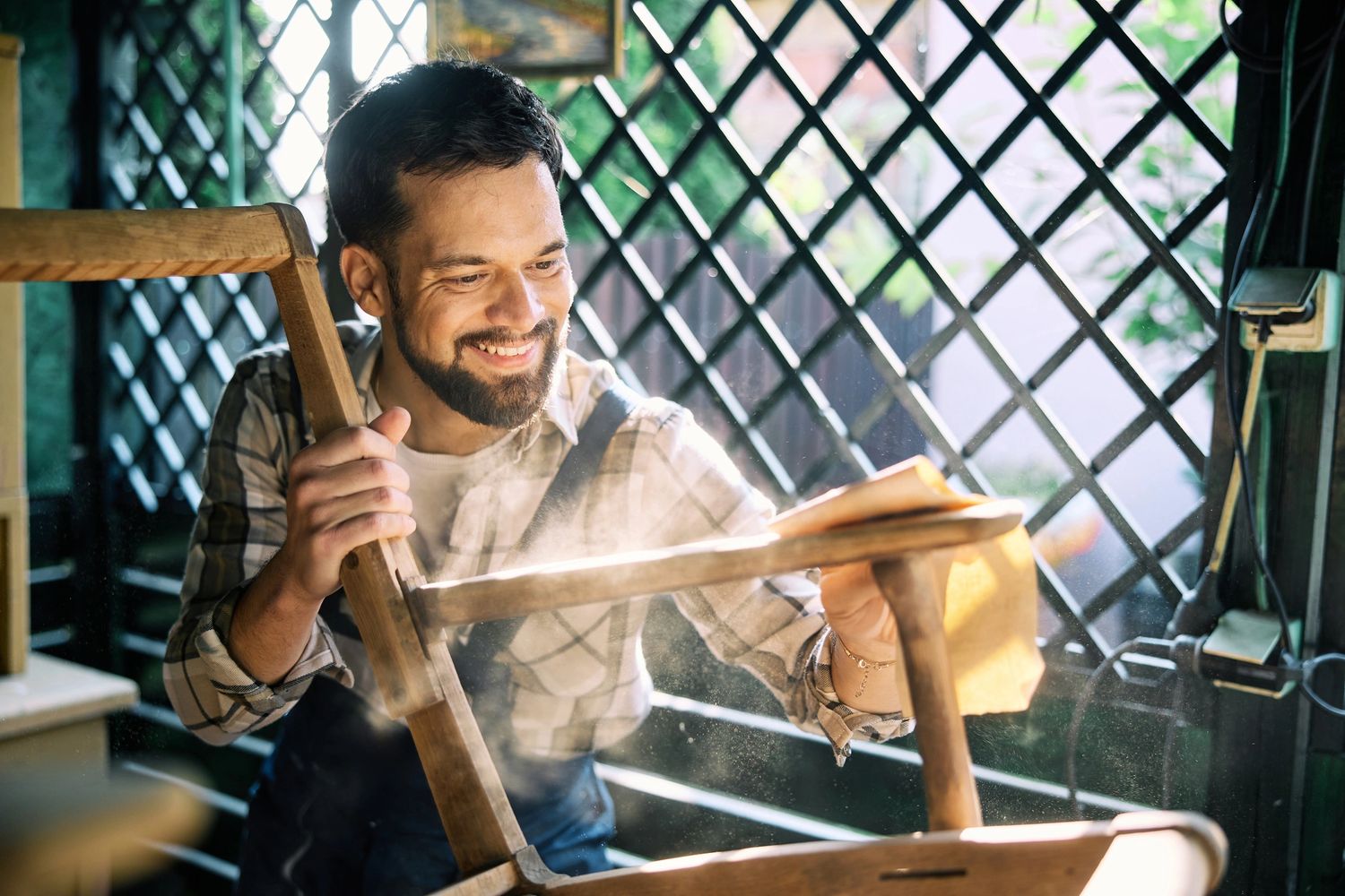 Man happily refurbishing a wooden chair with sandpaper in bright workshop.