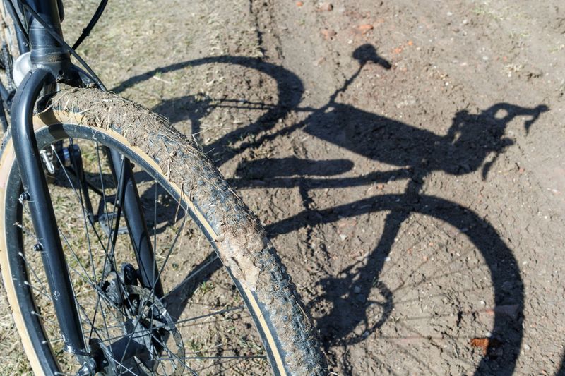 Close-up of a bicycle wheel covered in mud, standing on a rugged dirt road in a rural setting with dry grass in the background.