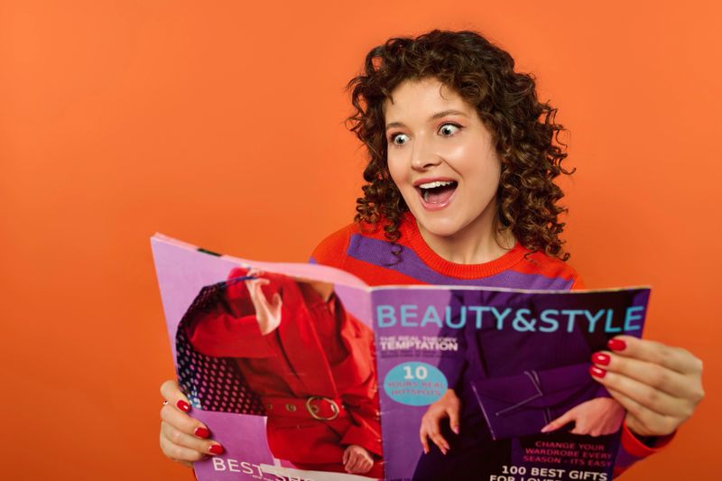 A young woman with curly hair joyfully browses a vibrant fashion magazine.
