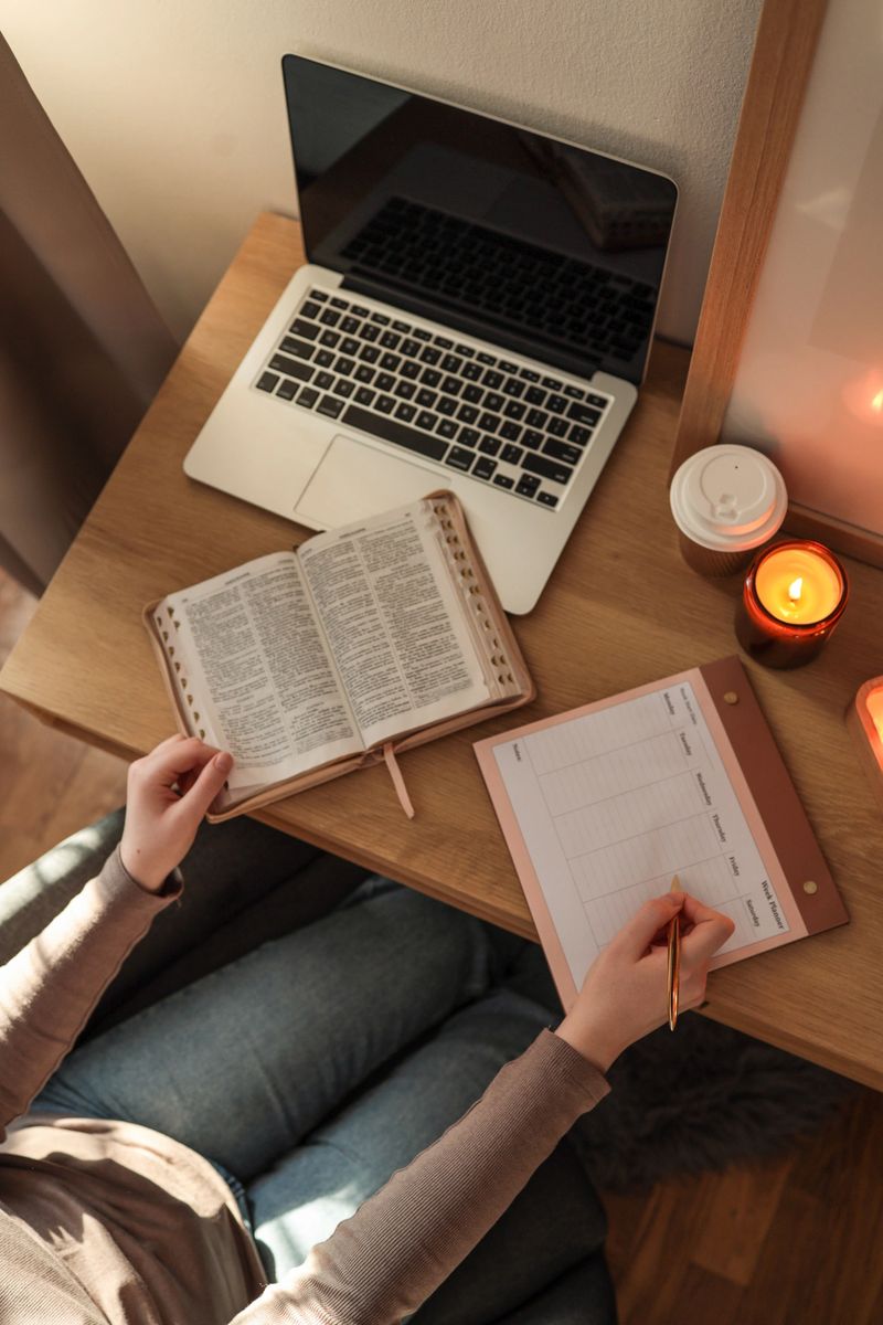 Person sitting at a wooden desk with an open Bible, a laptop, and a weekly planner, writing with a gold pen, accompanied by lit candles and a takeaway coffee cup in a cozy, sunlit workspace.