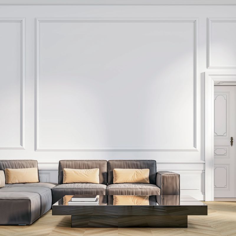 This image shows a sophisticated living room with classic wall paneling painted in white, creating a refined and elegant backdrop. A sleek dark gray sectional sofa with light beige cushions sits in front of the wall, paired with a modern, geometric black coffee table holding a few books. The space blends traditional architectural details with contemporary furniture for a stylish, balanced look. 3D rendered image.
