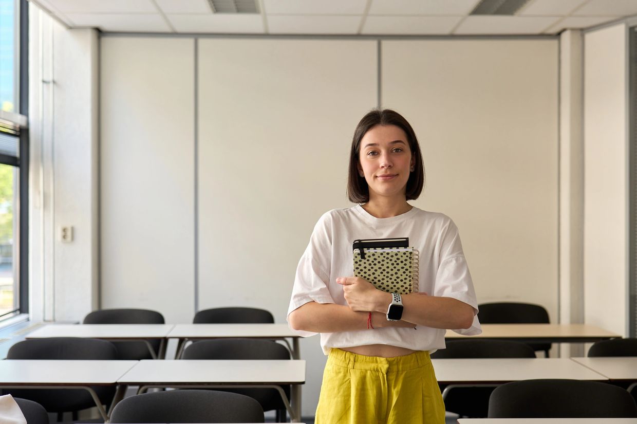 Young woman stands confidently in a bright classroom holding notebooks.