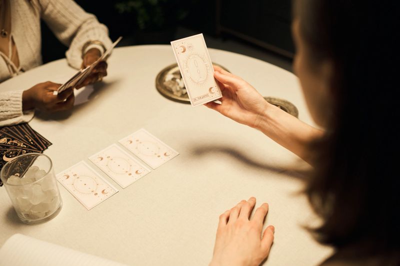 Caucasian young adult woman and Black female mystic sitting at table reading tarot cards, holding and examining cards with hands visible, engaging in fortune telling activity