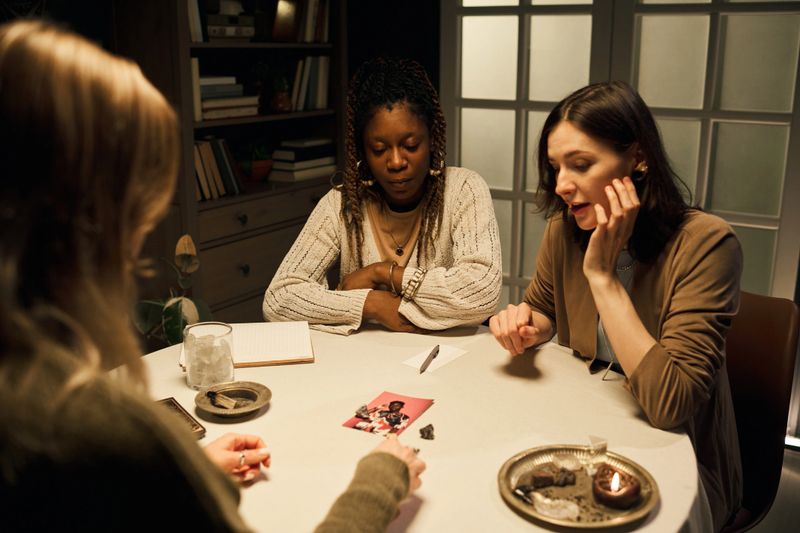 Two young women, one Black and one Caucasian, sitting at round table, receiving tarot card reading for love and relationships, discussing boyfriend photograph during intimate conversation