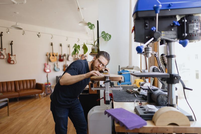 Luthier smoothing wooden body of electric guitar in his workshop, with other guitars hanging on the wall