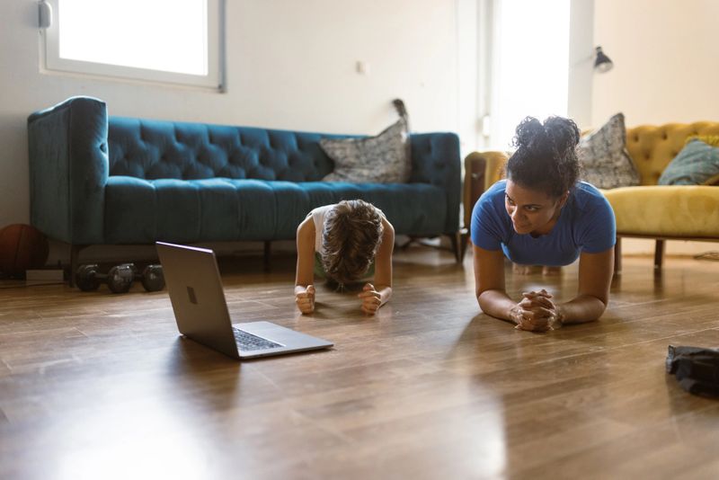 A mother and her child are in a plank position on the floor, following a workout routine from a laptop in a cozy living room setting.