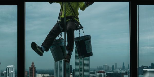 A worker suspended outside a building with buckets, cleaning windows high above a city.